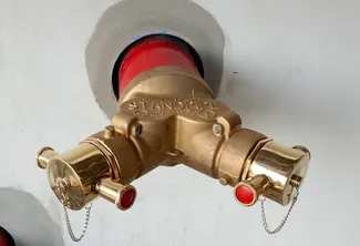 Brass dual-handle fire department connection valve mounted on a white wall, featuring two outlets with red caps and small chains attached